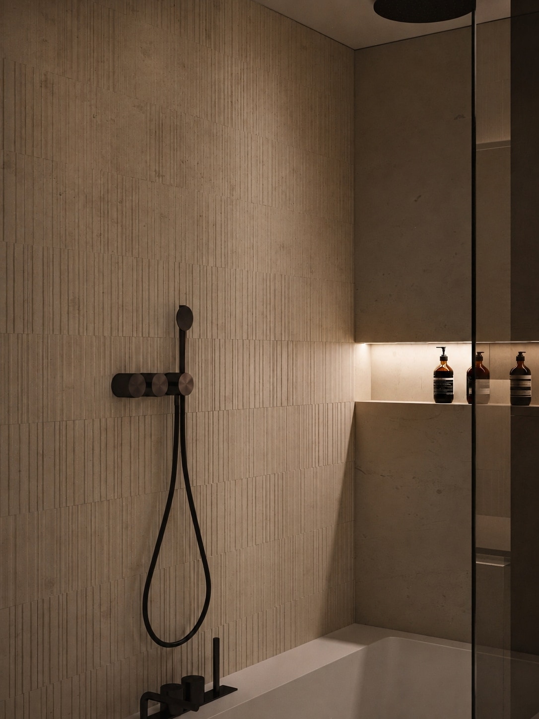 A matt bronze thermostatic shower valve and handset against fluted stone tiling, beside a backlit niche holding bottles.
