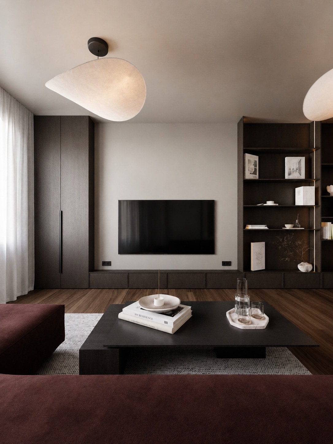 A living room with a dark joinery media wall, open display shelving, two organic pendant lights and an oxblood sofa.
