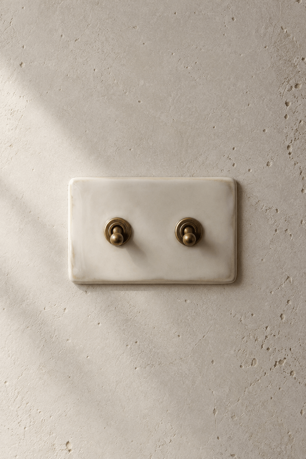A bone-glazed ceramic twin toggle with aged brass hardware on a pale mineral wall.